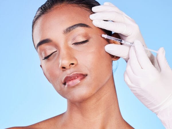 Belle Ange Health Service Ajax Needle, woman and face in studio for skincare, beauty process and aesthetic filler on blue background. Indian model, hands of surgeon and injection for plastic surgery, facial change or prp cosmetics